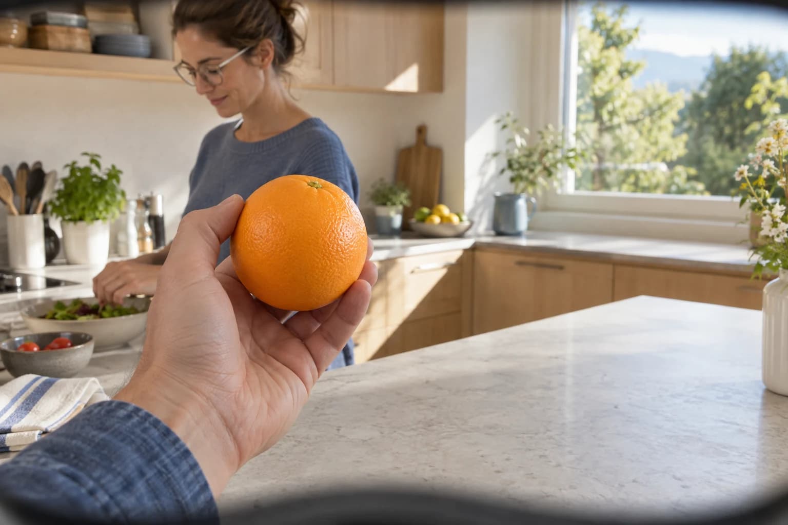 Aus der Brillenperspektive hält eine Hand eine Orange in einer hellen Küche; im Hintergrund kocht eine Person mit Brille.